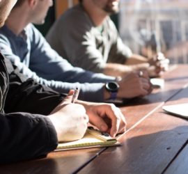 person writing at a desk in conversation with coworker