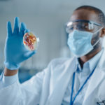 African American Male Scientist Wearing Face Mask and Glasses Looking at Petri Dish with Genetically Modified Sample Chemicals. Microbiologist Working in Modern Laboratory with Technological Equipment