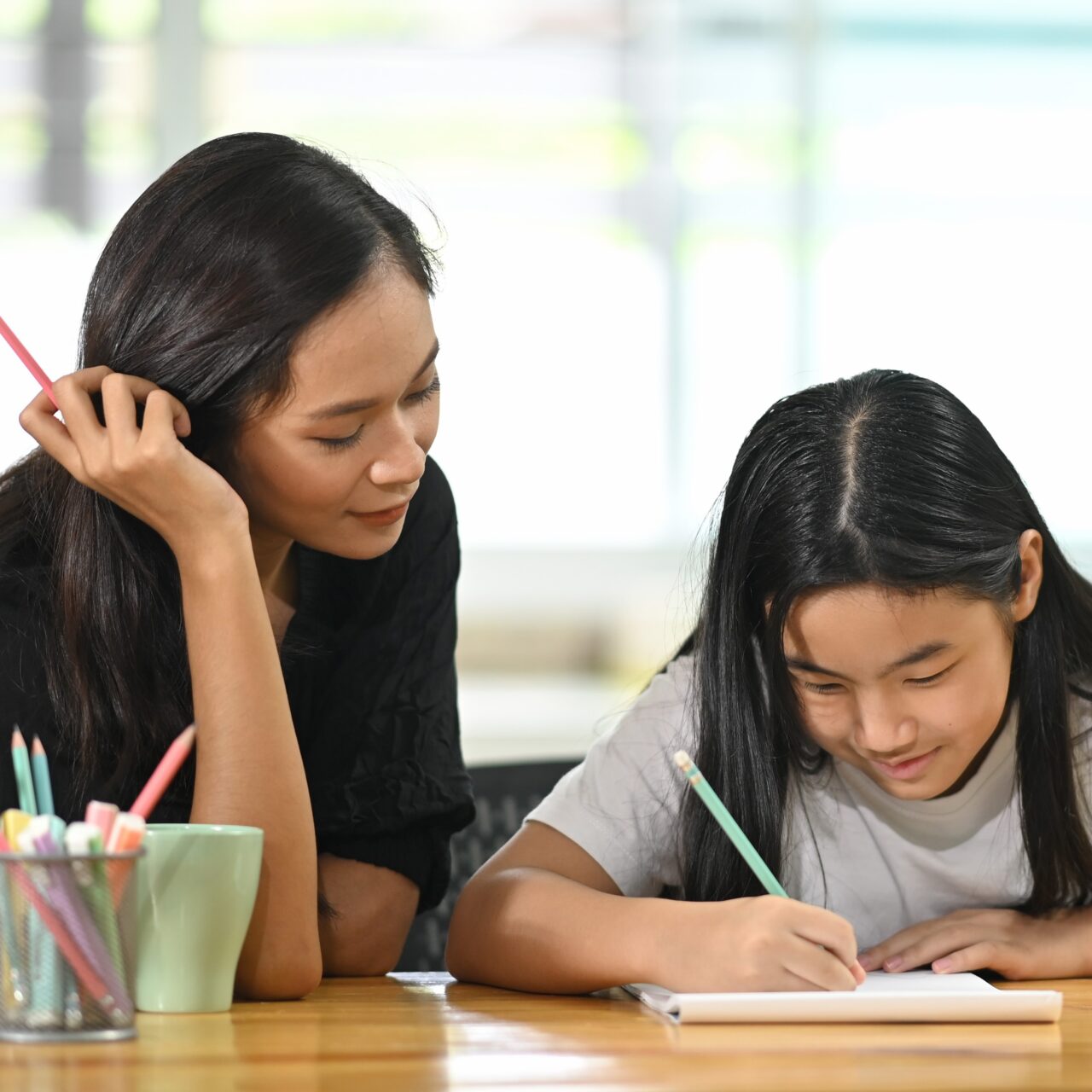 A young mother is teaching a daughter to do homework at the wooden table.