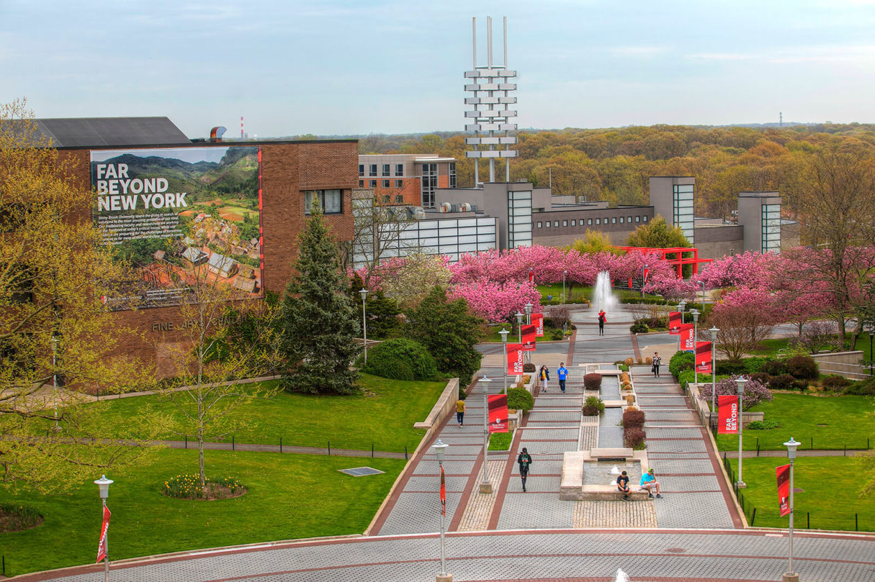 A photo of Stony Brook University's campus, lush with trees blooming flowers, green space, modern buildings, and a fountain in the middle of a circle pavillion
