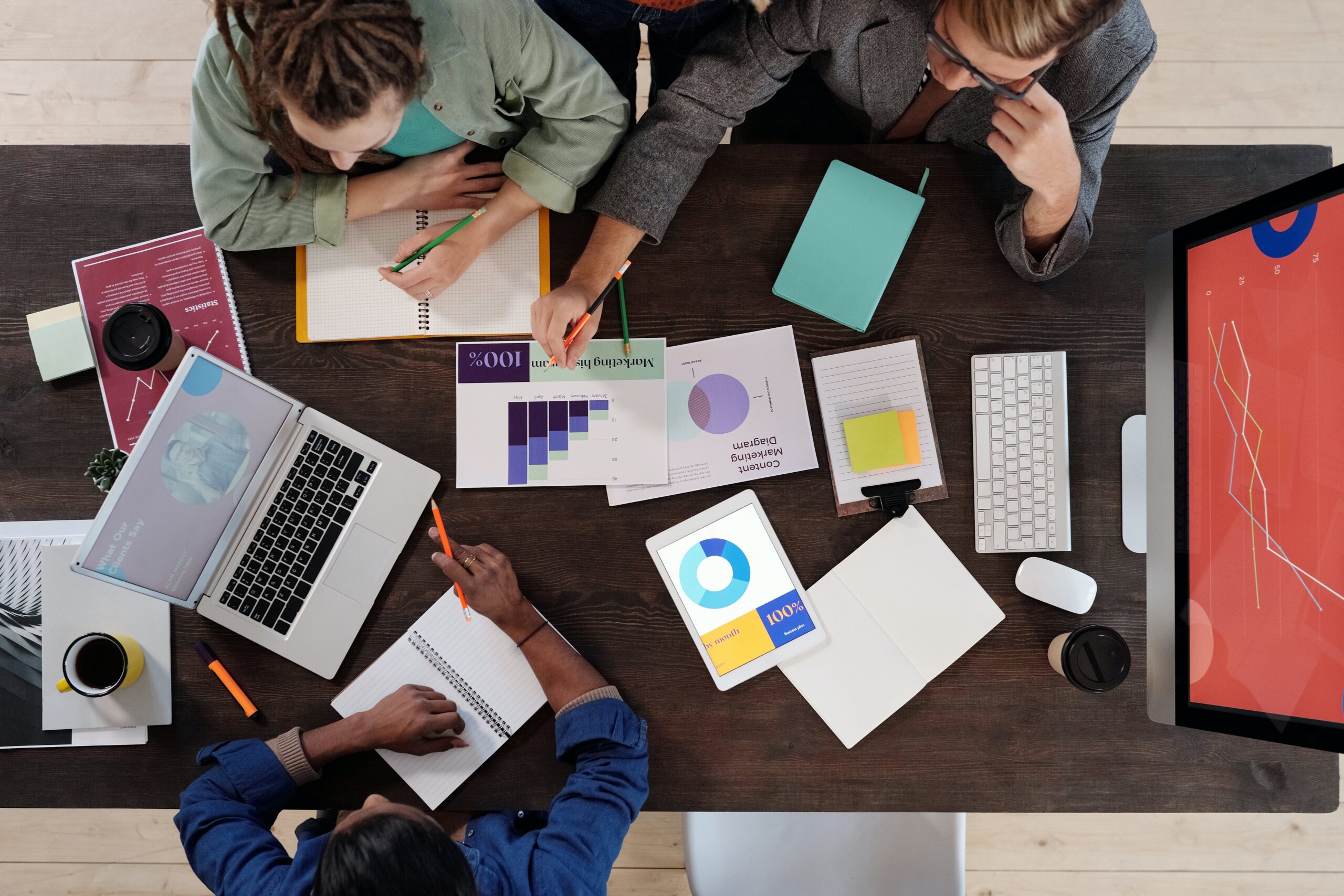 Overhead view of students together at a large wooden table, reviewing a set of printed graphs and computer dashboards of data